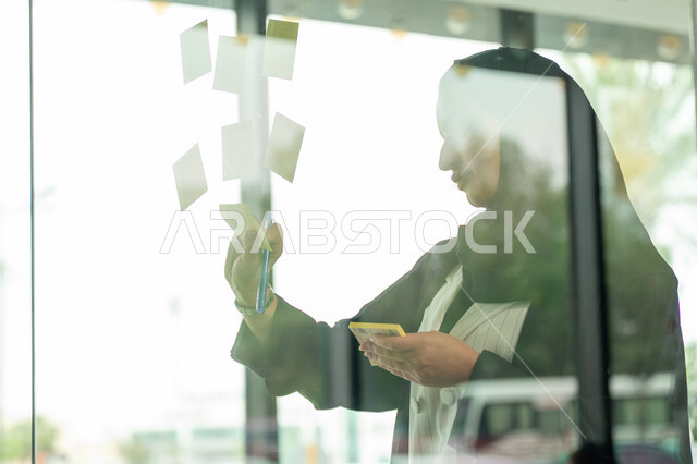 كتابة وتدوين شيء ما, أوراق الملاحظات الملونة المعلقة, بيئة العمل في الشركات السعودية, مهن وأعمال مكتبية, صورة مقربة لامرأة عربية خليجية سعودية ترتدي الحجاب والعباءة تقوم بلصق ورقة ملاحظة على واجهة زجاجية داخل المكتب