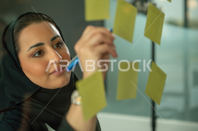 كتابة وتدوين شيء ما, أوراق الملاحظات الملونة المعلقة, بيئة العمل في الشركات السعودية, مهن وأعمال مكتبية, صورة مقربة لامرأة عربية خليجية سعودية ترتدي الحجاب والعباءة تقوم بلصق ورقة ملاحظة على واجهة زجاجية داخل المكتب