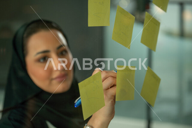 Writing and taking notes, colorful sticky notes, work environment in Saudi companies, office jobs and professions, close-up of a Saudi Arabian Gulf woman wearing a hijab and abaya sticking a note on a glass front inside the office