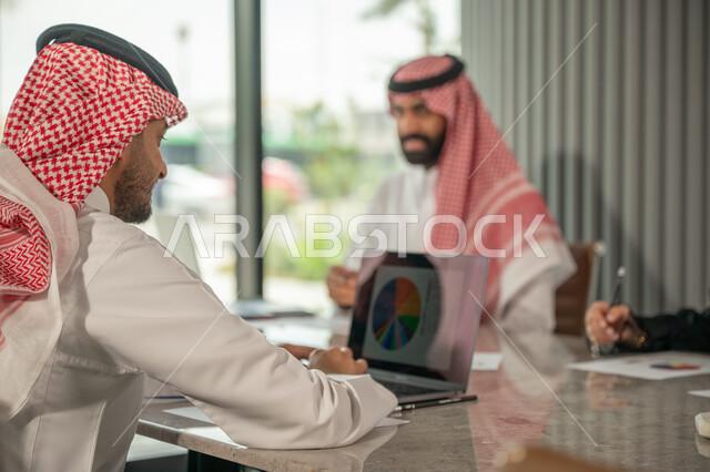 Explaining and analyzing graphs, integrating modern technical devices into office work, studying statistical data sheets, developing projects and investments, managing and dividing tasks, an Arab Gulf Saudi team conducting a business meeting in the conference room at the company's headquarters
