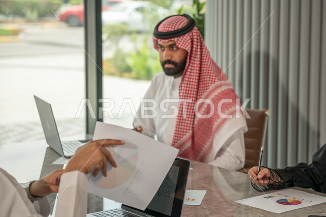 Explaining and analyzing graphs, integrating modern technical devices into office work, studying statistical data sheets, developing projects and investments, managing and dividing tasks, an Arab Gulf Saudi team conducting a business meeting in the conference room at the company's headquarters