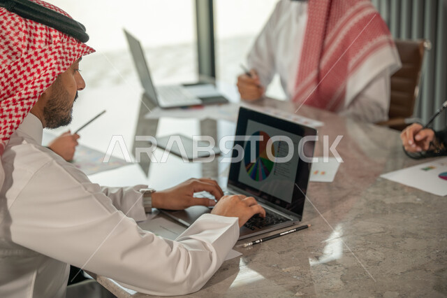 Explaining and analyzing graphs, integrating modern technical devices into office work, studying statistical data sheets, developing projects and investments, managing and dividing tasks, an Arab Gulf Saudi team conducting a business meeting in the conference room at the company's headquarters