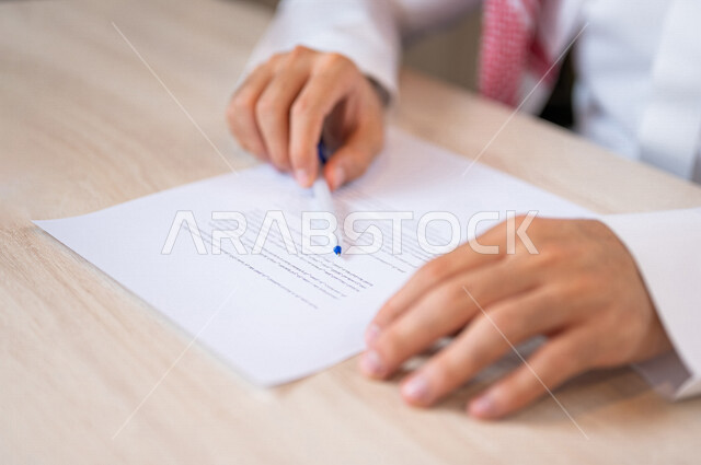 رجل عربي خليجي سعودي امامه أوراق خاصة بعقد وفوقها قلم، سكرتير يدير الاعمال الورقية، قراءة البنود الخاصة بالعقد، التحقق والتدقيق في شروط العقد، الموافقة على الشروط