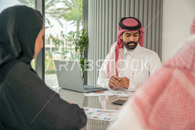 Explaining and analyzing graphs, integrating modern technical devices into office work, studying statistical data sheets, developing projects and investments, managing and dividing tasks, an Arab Gulf Saudi team conducting a business meeting in the conference room at the company's headquarters