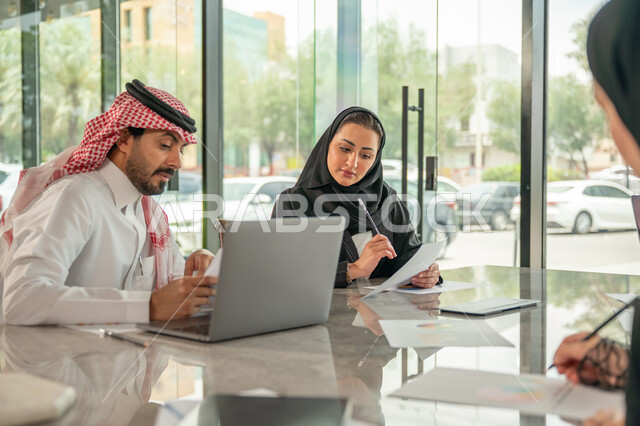 Explaining and analyzing graphs, integrating modern technical devices into office work, studying statistical data sheets, developing projects and investments, managing and dividing tasks, an Arab Gulf Saudi team conducting a business meeting in the conference room at the company's headquarters