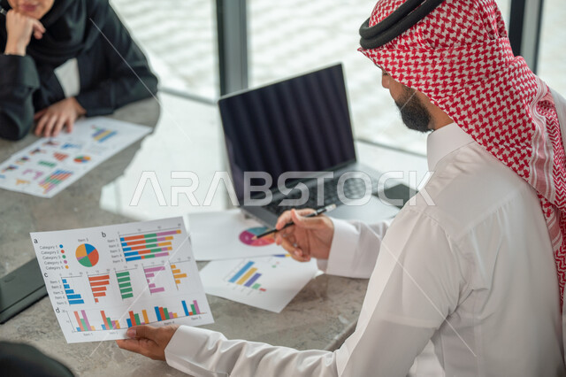 Explaining and analyzing graphs, integrating modern technical devices into office work, studying statistical data sheets, developing projects and investments, managing and dividing tasks, an Arab Gulf Saudi team conducting a business meeting in the conference room at the company's headquarters