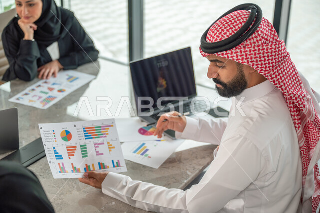 Explaining and analyzing graphs, integrating modern technical devices into office work, studying statistical data sheets, developing projects and investments, managing and dividing tasks, an Arab Gulf Saudi team conducting a business meeting in the conference room at the company's headquarters