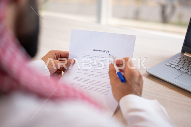 رجل عربي خليجي سعودي يجلس امام الحاسوب المحمول ويمسك بيده قلم ويقوم بقراءة وثيقة، سكرتير يدير الاعمال الورقية، التحقق والتدقيق في الوثيقة، مفهوم التوظيف والعمل، أعمال مكتبية