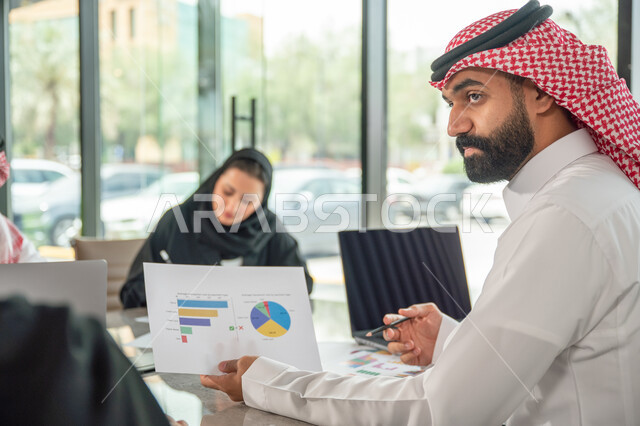 Explaining and analyzing graphs, integrating modern technical devices into office work, studying statistical data sheets, developing projects and investments, managing and dividing tasks, an Arab Gulf Saudi team conducting a business meeting in the conference room at the company's headquarters