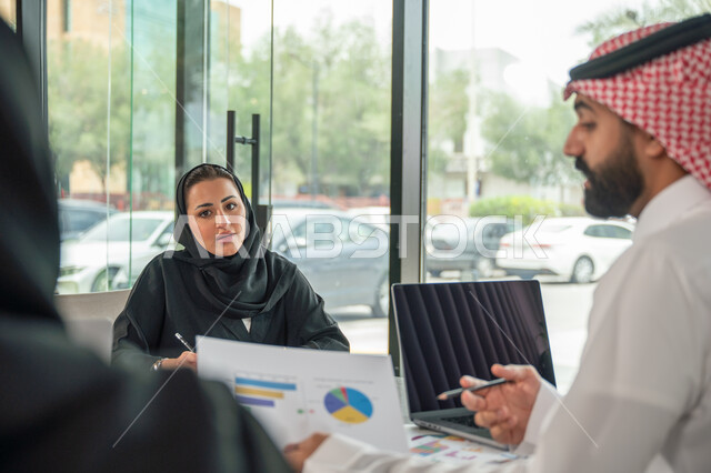 Explaining and analyzing graphs, integrating modern technical devices into office work, studying statistical data sheets, developing projects and investments, managing and dividing tasks, an Arab Gulf Saudi team conducting a business meeting in the conference room at the company's headquarters