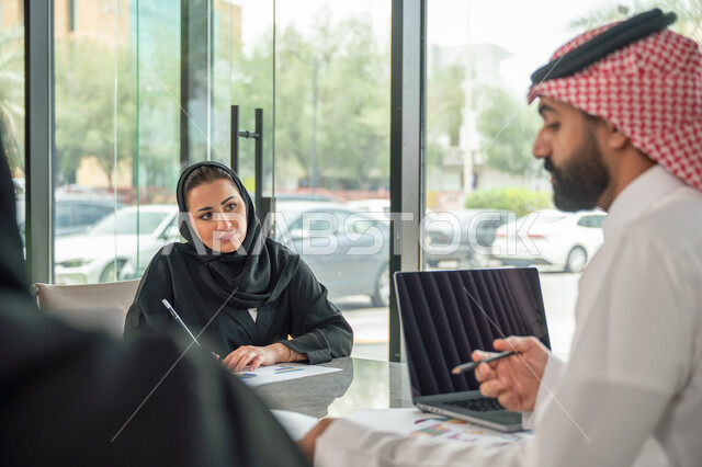 Explaining and analyzing graphs, integrating modern technical devices into office work, studying statistical data sheets, developing projects and investments, managing and dividing tasks, an Arab Gulf Saudi team conducting a business meeting in the conference room at the company's headquarters