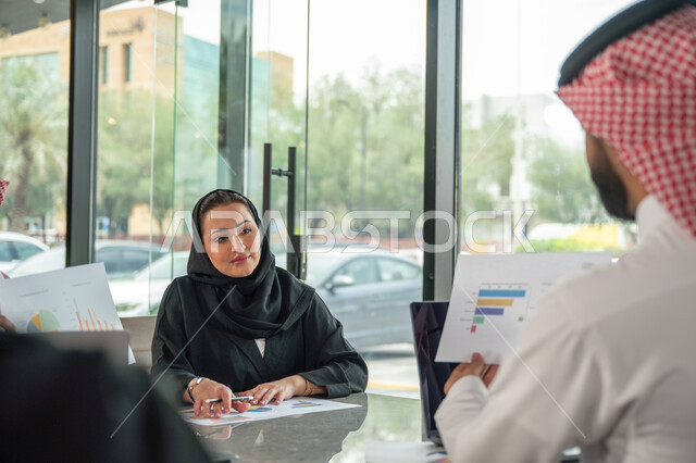 Explaining and analyzing graphs, integrating modern technical devices into office work, studying statistical data sheets, developing projects and investments, managing and dividing tasks, an Arab Gulf Saudi team conducting a business meeting in the conference room at the company's headquarters