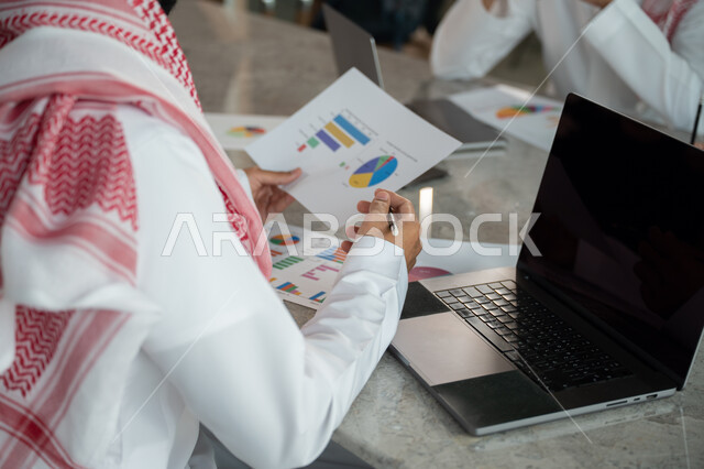 Explaining and analyzing graphs, integrating modern technical devices into office work, studying statistical data sheets, developing projects and investments, managing and dividing tasks, an Arab Gulf Saudi team conducting a business meeting in the conference room at the company's headquarters
