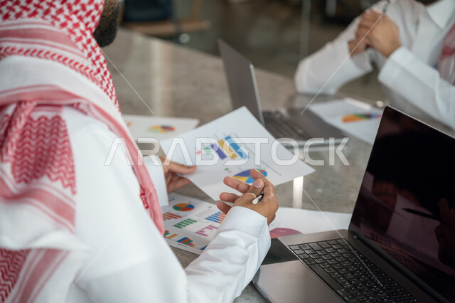 Explaining and analyzing graphs, integrating modern technical devices into office work, studying statistical data sheets, developing projects and investments, managing and dividing tasks, an Arab Gulf Saudi team conducting a business meeting in the conference room at the company's headquarters