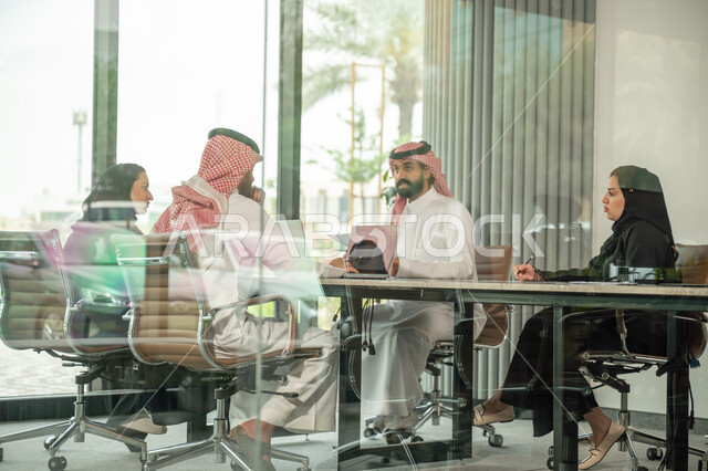 Explaining and analyzing graphs, integrating modern technical devices into office work, studying statistical data sheets, developing projects and investments, managing and dividing tasks, an Arab Gulf Saudi team conducting a business meeting in the conference room at the company's headquarters