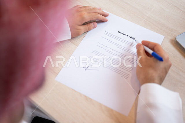 رجل عربي خليجي سعودي يمسك بيده قلم ويقوم بقراءة وثيقة، سكرتير يدير الاعمال الورقية، التحقق والتدقيق في الوثيقة، مفهوم التوظيف والعمل، أعمال مكتبية