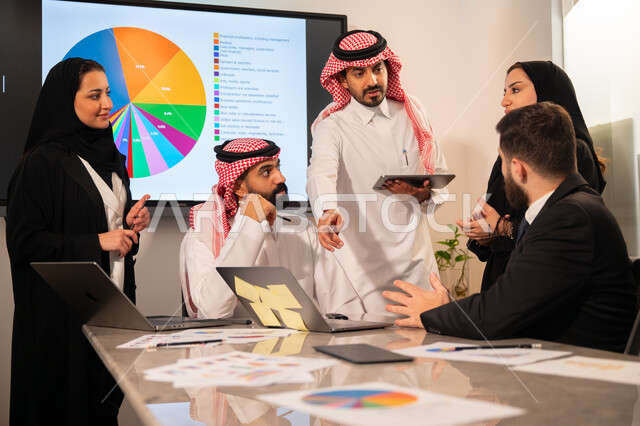 دمج الأجهزة التقنية الحديثة بالأعمال المكتبية، دراسة أوراق البيانات الإحصائية, تطوير المشاريع و الاستثمارات, إدارة و تقسيم المهام، فريق عمل عربي خليجي سعودي يقوم بإجراء اجتماع عمل داخل غرفة الاجتماعات في مقر الشركة، شرح و تحليل الرسوم البيانية