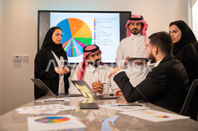 Integrating modern technical devices into office work, studying statistical data sheets, developing projects and investments, managing and dividing tasks, an Arab Gulf Saudi team conducting a business meeting in the company's headquarters meeting room, explaining and analyzing graphs.