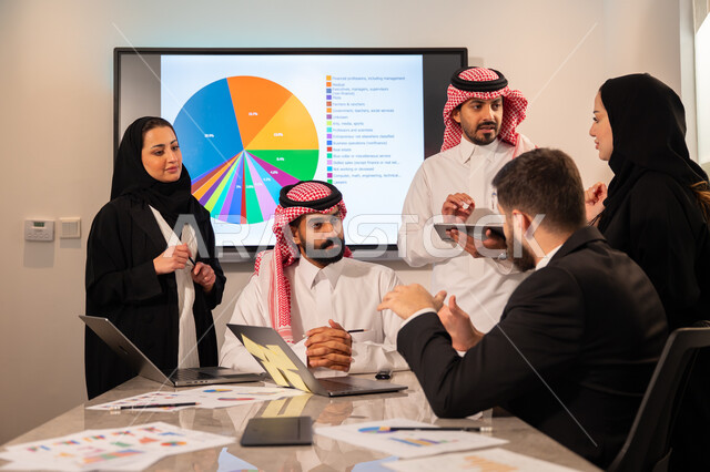 Integrating modern technical devices into office work, studying statistical data sheets, developing projects and investments, managing and dividing tasks, an Arab Gulf Saudi team conducting a business meeting in the company's headquarters meeting room, explaining and analyzing graphs.