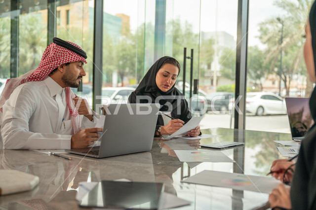 Integrating modern technical devices into office work, studying statistical data sheets, developing projects and investments, managing and dividing tasks, an Arab Gulf Saudi team conducting a business meeting in the company's headquarters meeting room, explaining and analyzing graphs.