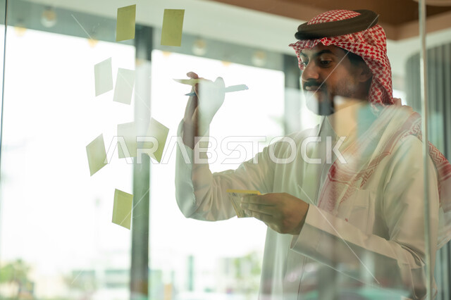 Writing and taking notes. Colorful sticky notes. Work environment in Saudi companies. Office jobs and professions. Close-up of a Saudi Arabian Gulf man wearing a traditional shemagh and thobe, sticking a note on a glass window inside the office.