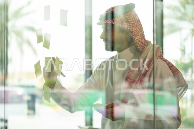 كتابة وتدوين شيء ما, أوراق الملاحظات الملونة المعلقة, بيئة العمل في الشركات السعودية, مهن وأعمال مكتبية, صورة مقربة لرجل عربي خليجي سعودي يرتدي الشماغ والثوب التقليدي يقوم بلصق ورقة ملاحظة على واجهة زجاجية داخل المكتب