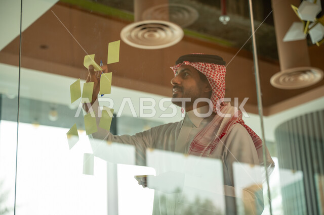 Writing and taking notes. Colorful sticky notes. Work environment in Saudi companies. Office jobs and professions. Close-up of a Saudi Arabian Gulf man wearing a traditional shemagh and thobe, sticking a note on a glass window inside the office.