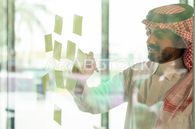 كتابة وتدوين شيء ما, أوراق الملاحظات الملونة المعلقة, بيئة العمل في الشركات السعودية, مهن وأعمال مكتبية, صورة مقربة لرجل عربي خليجي سعودي يرتدي الشماغ والثوب التقليدي يقوم بلصق ورقة ملاحظة على واجهة زجاجية داخل المكتب