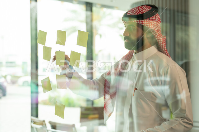 Writing and taking notes. Colorful sticky notes. Work environment in Saudi companies. Office jobs and professions. Close-up of a Saudi Arabian Gulf man wearing a traditional shemagh and thobe, sticking a note on a glass window inside the office.