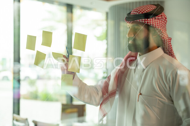 كتابة وتدوين شيء ما, أوراق الملاحظات الملونة المعلقة, بيئة العمل في الشركات السعودية, مهن وأعمال مكتبية, صورة مقربة لرجل عربي خليجي سعودي يرتدي الشماغ والثوب التقليدي يقوم بلصق ورقة ملاحظة على واجهة زجاجية داخل المكتب