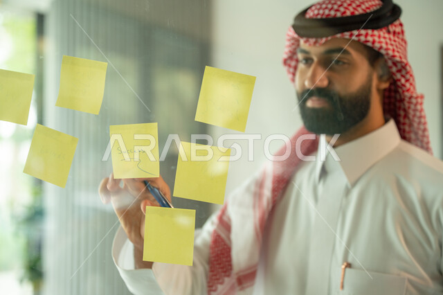 Writing and taking notes. Colorful sticky notes. Work environment in Saudi companies. Office jobs and professions. Close-up of a Saudi Arabian Gulf man wearing a traditional shemagh and thobe, sticking a note on a glass window inside the office.