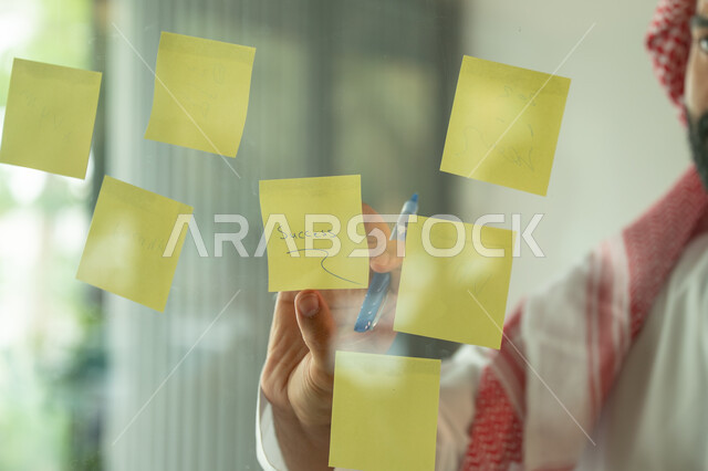 Writing and taking notes. Colorful sticky notes. Work environment in Saudi companies. Office jobs and professions. Close-up of a Saudi Arabian Gulf man wearing a traditional shemagh and thobe, sticking a note on a glass window inside the office.