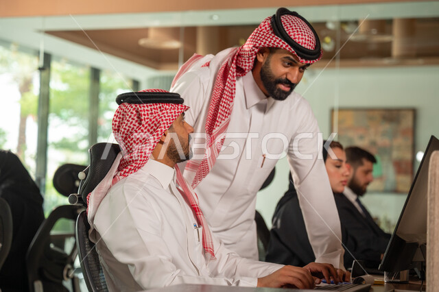 استخدام الحاسب والتقنية في مجال الاعمال، بيئة العمل المريحة في الشركات السعودية، الحوار و النقاش بين الزملاء، تبادل الخبرات والمعلومات لتحقيق الاهداف, موظفان عربيان خليجيان سعوديان يجلسان على مكتبهم داخل مقر الشركة