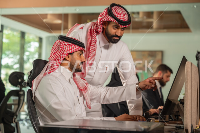  استخدام الحاسب والتقنية في مجال الاعمال، بيئة العمل المريحة في الشركات السعودية، الحوار و النقاش بين الزملاء، تبادل الخبرات والمعلومات لتحقيق الاهداف, موظفان عربيان خليجيان سعوديان يجلسان على مكتبهم داخل مقر الشركة