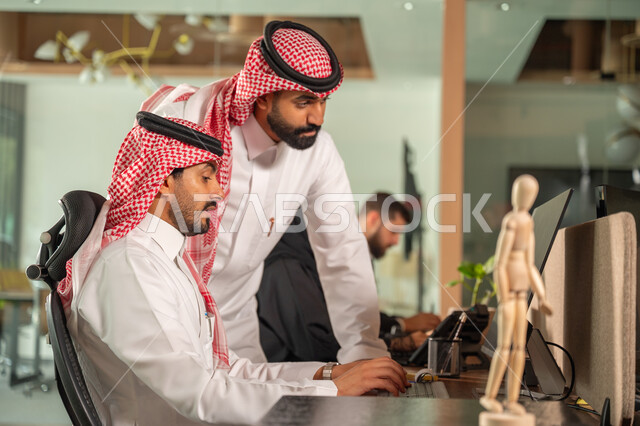  استخدام الحاسب والتقنية في مجال الاعمال، بيئة العمل المريحة في الشركات السعودية، الحوار و النقاش بين الزملاء، تبادل الخبرات والمعلومات لتحقيق الاهداف, موظفان عربيان خليجيان سعوديان يجلسان على مكتبهم داخل مقر الشركة
