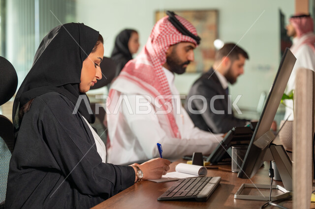 Using computers and technology in business, comfortable work environment in Saudi companies, dialogue and discussion between colleagues, exchange of experiences and information to achieve goals, two Saudi Gulf Arab employees sitting at their desk inside the company headquarters