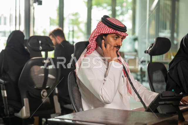 Communicating with customers by phone, a side view of a Saudi Arabian Gulf employee wearing a traditional thobe and shemagh working at the company headquarters, completing daily work tasks, Saudi professions and jobs, a comfortable work environment in Saudi companies.