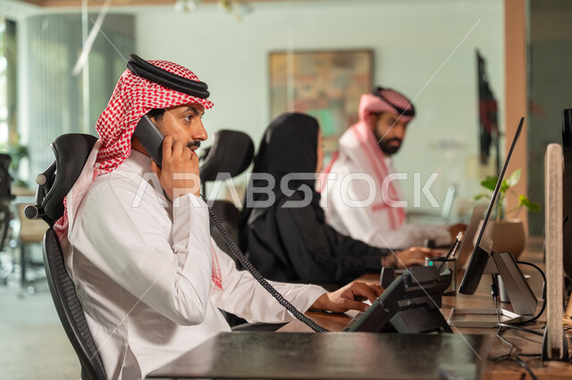 Communicating with customers by phone, a side view of a Saudi Arabian Gulf employee wearing a traditional thobe and shemagh working at the company headquarters, completing daily work tasks, Saudi professions and jobs, a comfortable work environment in Saudi companies.