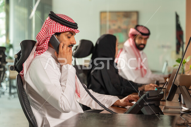  التواصل مع العملاء من خلال الهاتف، صورة من الجانب لموظف عربي سعودي خليجي يرتدي الثوب التقليدي والشماغ ويعمل في مقر الشركة، انجاز مهام العمل اليومية، مهن ووظائف سعودية، بيئة العمل المريحة في الشركات السعودية