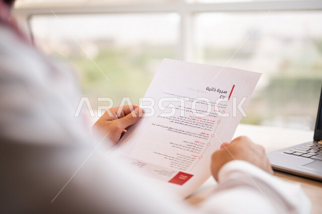 رجل عربي خليجي سعودي يجلس أمام الحاسوب المحمول ويحمل بيده اوراق خاصة بالسيرة الذاتية، سكرتير يدير الاعمال الورقية، قراءة السيرة الذاتية، مفهوم التوظيف والعمل، أعمال مكتبية