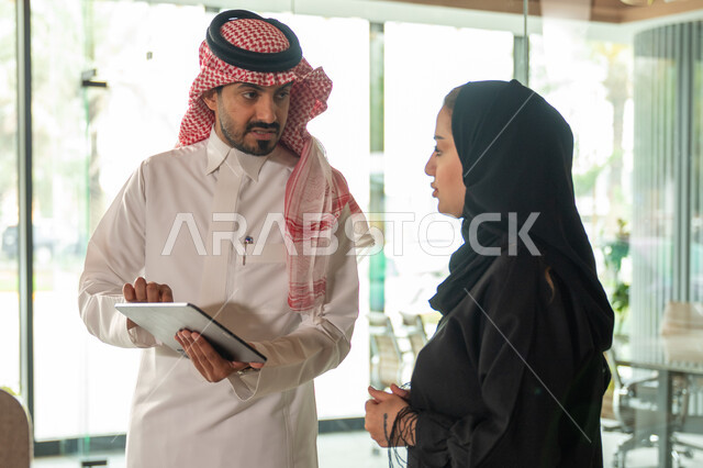 Discussing and distributing work tasks, the Saudi work environment, work development and problem solving, Saudi office professions and jobs, exchanging opinions and experiences, a meeting of a Saudi Gulf Arab businessman and woman at the company headquarters, managing work using a technical device, unity and teamwork, achieving the company's goals.