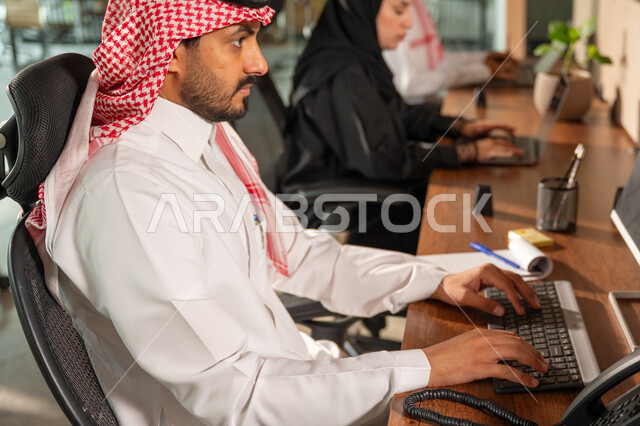 Gestures and expressions of concentration, Saudi office jobs and professions, writing and recording data and results, managing and completing tasks using a modern and advanced technical device, a close-up side view of a Saudi Arabian Gulf man wearing a traditional shemagh and thobe working on a laptop