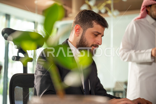 ادارة وانجاز المهام باستخدام جهاز تقني حديث ومتطور, صورة مقربة من الجانب لرجل عربي خليجي سعودي يرتدي البدلة الرسمية يعمل على الحاسوب المحمول، ايماءات وتعابير التركيز، مهن ووظائف مكتبية سعودية، كتابة وتدوين البيانات والنتائج