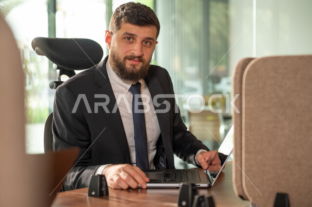 ادارة وانجاز المهام باستخدام جهاز تقني حديث ومتطور, صورة مقربة من الجانب لرجل عربي خليجي سعودي يرتدي البدلة الرسمية يعمل على الحاسوب المحمول، ايماءات وتعابير التركيز، مهن ووظائف مكتبية سعودية، كتابة وتدوين البيانات والنتائج