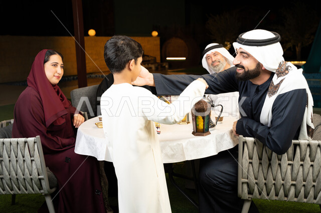 ديكور وزينة المناسبات والأعياد الاسلامية، اجتماع العائلة في شهر رمضان المبارك، أسرة عربية خليجية إماراتية تقضي وقت ممتع في جلسة خارجية في احدى الشاليهات، صورة من الخلف لولد يحمل بيده فانوس معدني مضيء