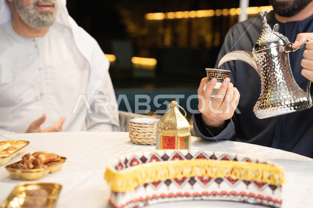 الاستمتاع بالمشروبات الساخنة, منتج محلي وطني، أصالة العادات والتقاليد الأصيلة, التمسك بالقيم الكريمة المتوارثة من الأجداد، كرم الضيافة وحسن الاستقبال, صورة مقربة ليد رجل عربي خليجي اماراتي يقوم بصب القهوة من الدلة الفضية، زينة وديكور شهر رمضان المبارك
