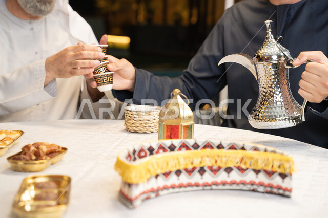 التمسك بالقيم الكريمة المتوارثة من الأجداد، أصالة العادات والتقاليد الأصيلة, الاستمتاع بالمشروبات الساخنة, منتج محلي وطني، كرم الضيافة وحسن الاستقبال, صورة مقربة ليد رجل عربي خليجي اماراتي يقوم بصب القهوة من الدلة الفضية، زينة وديكور شهر رمضان المبارك