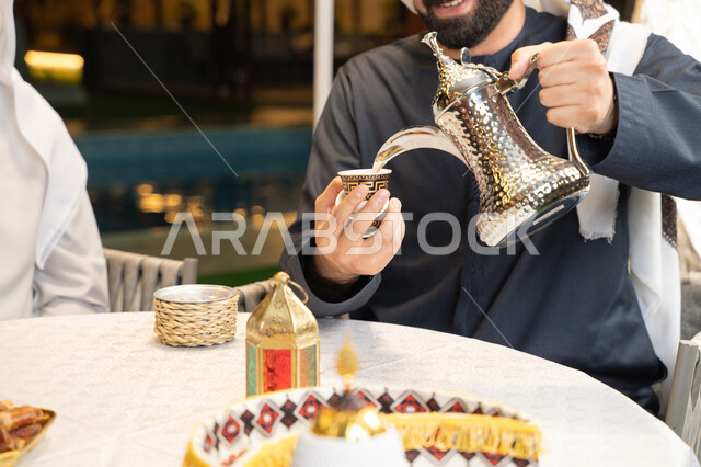 التمسك بالقيم الكريمة المتوارثة من الأجداد، أصالة العادات والتقاليد الأصيلة, الاستمتاع بالمشروبات الساخنة, منتج محلي وطني، كرم الضيافة وحسن الاستقبال, صورة مقربة ليد رجل عربي خليجي اماراتي يقوم بصب القهوة من الدلة الفضية، زينة وديكور شهر رمضان المبارك