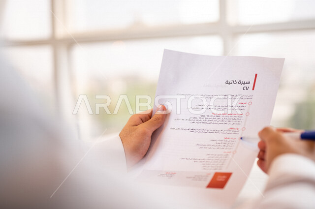 رجل عربي خليجي سعودي يقوم بقراءة اوراق خاصة بالسيرة الذاتية، سكرتير يدير الاعمال الورقية، قراءة السيرة الذاتية، مفهوم التوظيف والعمل، أعمال مكتبية