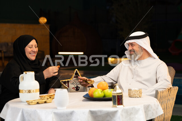 قضاء وقت ممتع في جلسة خارجية في احدى الشاليهات، زينة وديكور المناسبات والأعياد الاسلامية، أجواء عائلية سعيدة في شهر رمضان المبارك، زوجان عربيان خليجيان اماراتيان يقومان بتناول التمربإيماءات السعادة والسرور